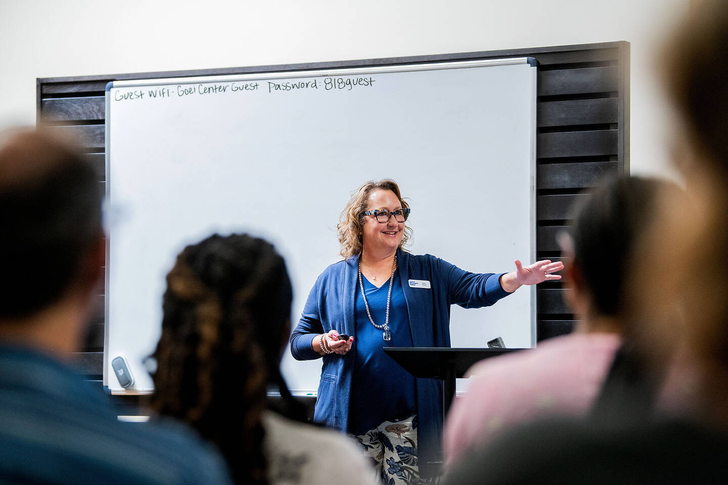 A person stands at a podium in front of an audience, gesturing while speaking, with a whiteboard behind them displaying guest Wi-Fi information.